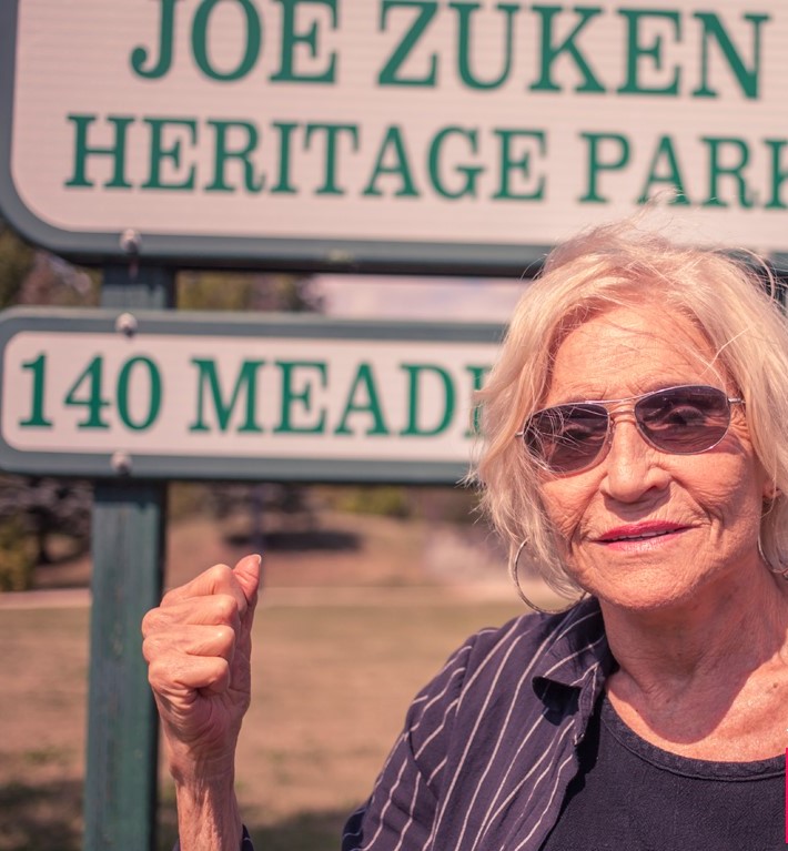 Fagie Fainman in a black shirt and a blue and white stripe button-up. She wears sunglasses with short white hair with her right fist raised. She stands in front of a park sign.