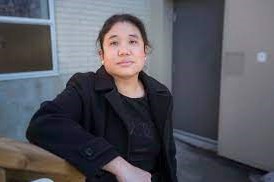 Vivienne Ho in a black shirt with a black jacket. Their hair is pulled back and their arm rests on a railing with a building behind them.